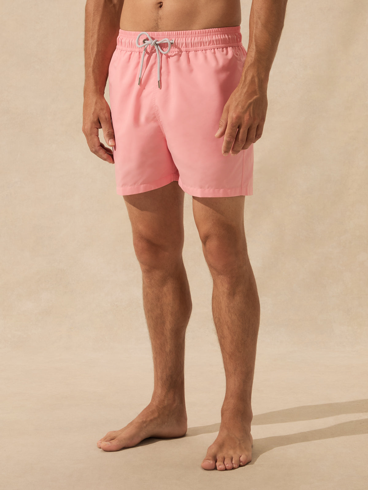 A man wearing light pink swim trunks with a white drawstring, standing barefoot against a warm beige textured studio backdrop.