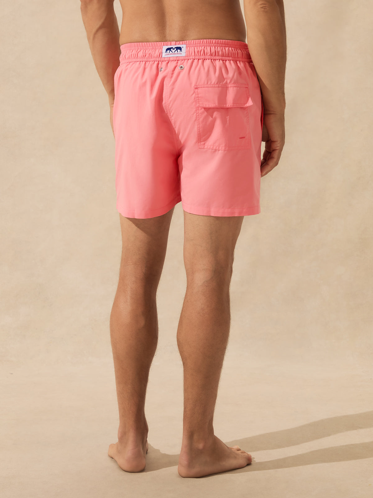 A man wearing coral pink swim trunks, shown from the back against a neutral beige background.