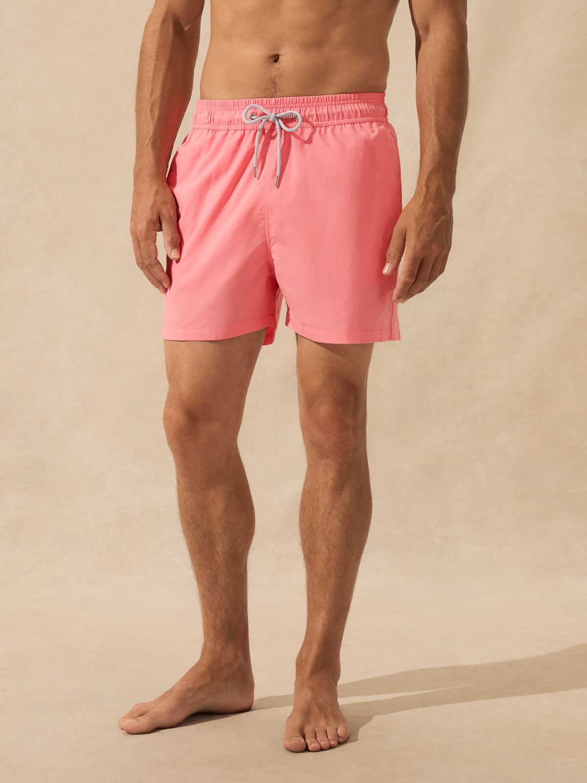 A man wearing bright pink swim trunks with a white drawstring, standing barefoot against a neutral beige background.
