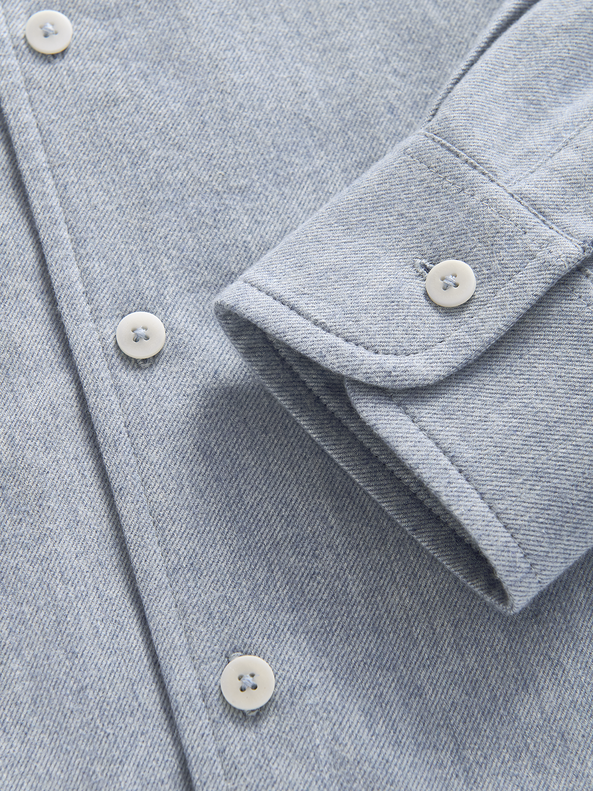 Close-up of a light blue textured fabric garment, showing part of a sleeve cuff with a button and the front placket with white buttons.