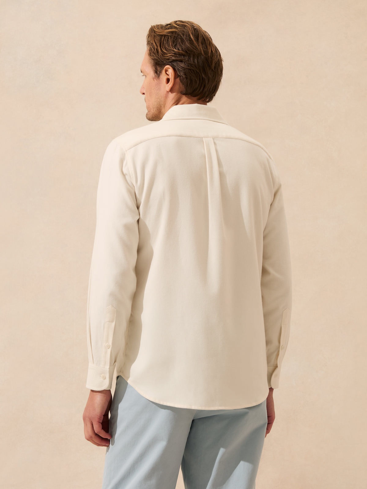 A man shown from the back wearing a cream-coloured long-sleeve button-down shirt and light blue trousers, standing against a textured beige background.