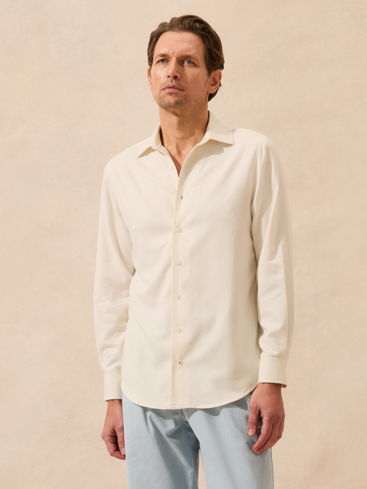 A man wearing a cream-coloured long-sleeve button-down shirt and light blue trousers, standing against a textured beige background.