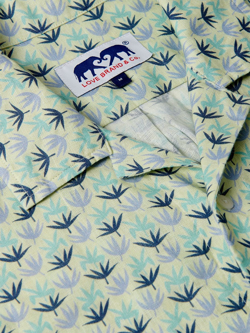 Close-up view of Men's Palm Sugar Arawak Linen Shirt featuring a tropical fan palm print. Men's linen shirt in sky blue, aqua green, and deep blue on a limoncello yellow base, with a Love Brand & Co. label.