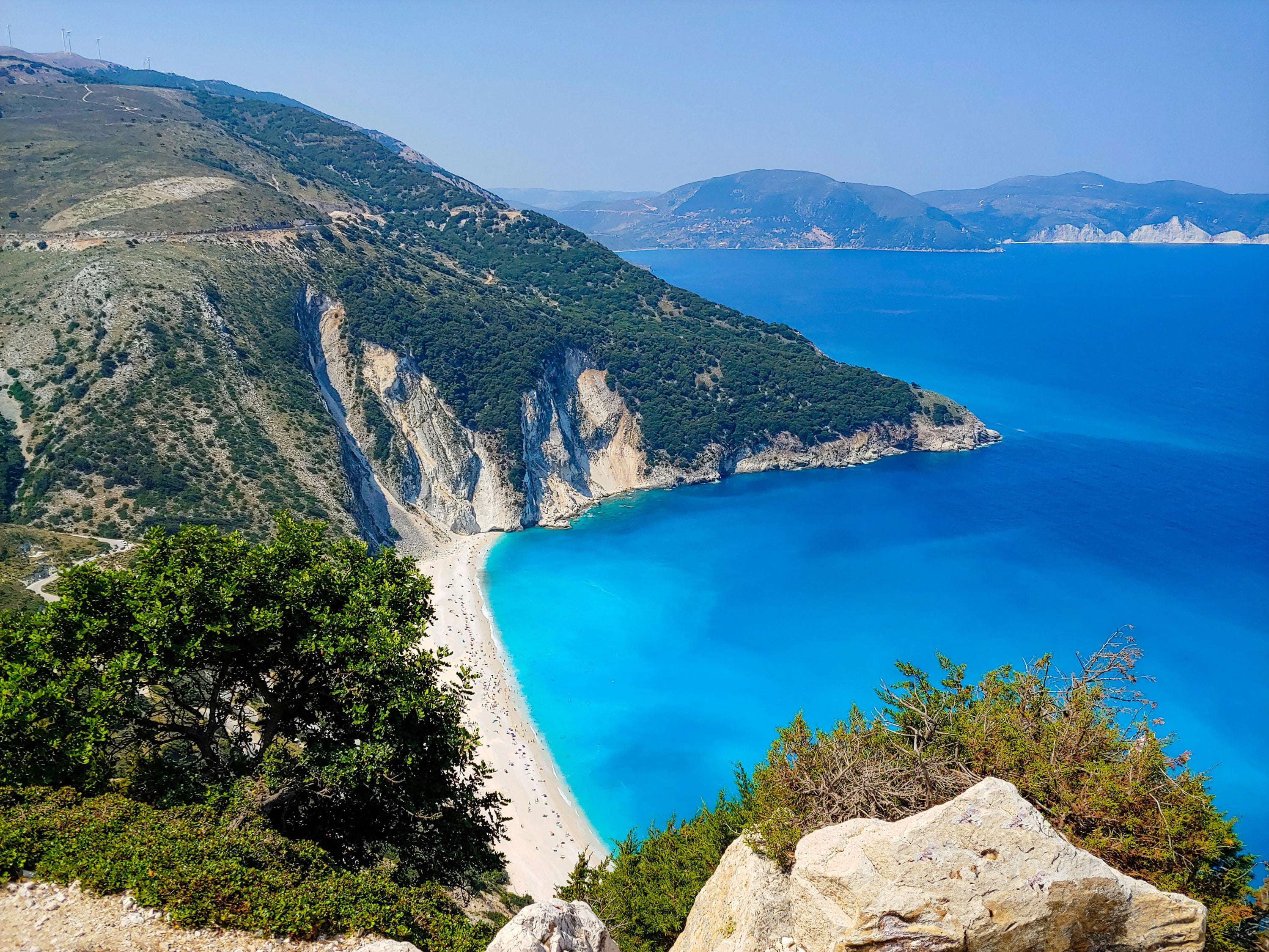 Coastal landscape with blue water, greenery, and mountains under a clear sky.