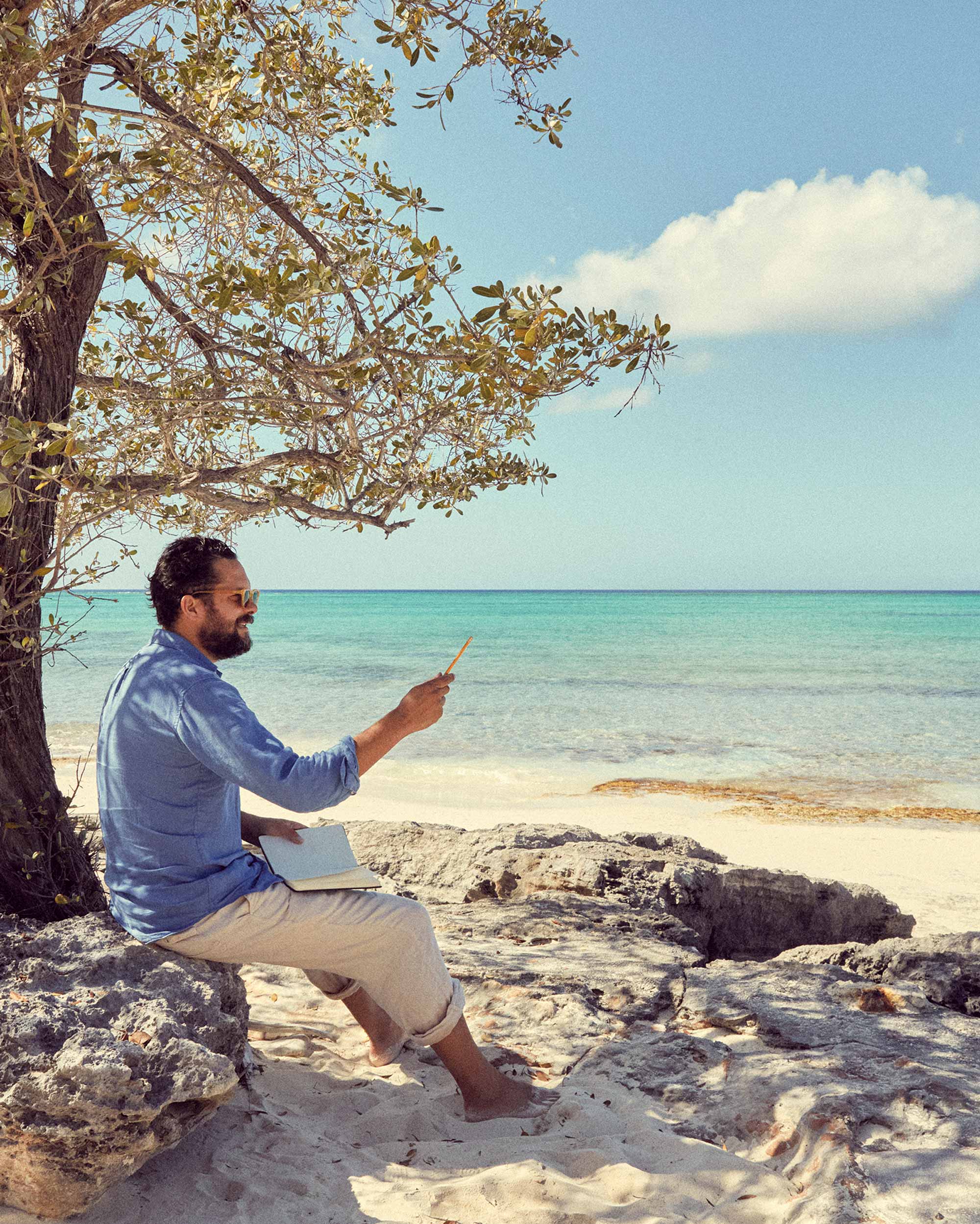 Oliver Tomalin sitting by a tree on a beach with ocean view sketching new prints