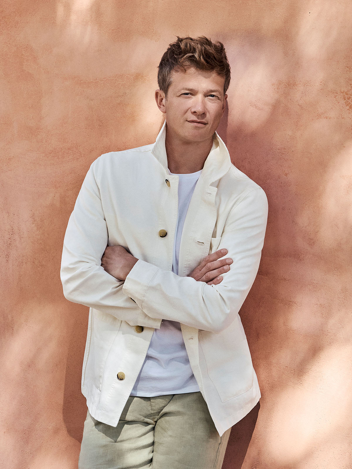 Ed Speleers wearing a cream-colored jacket over a white t-shirt and khaki pants, standing with arms crossed against a textured terracotta wall.