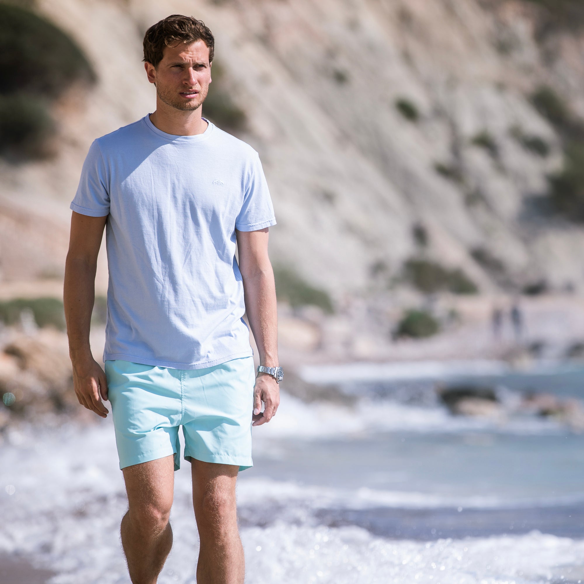 Man wearing a light blue t-shirt and shorts standing on a beach.