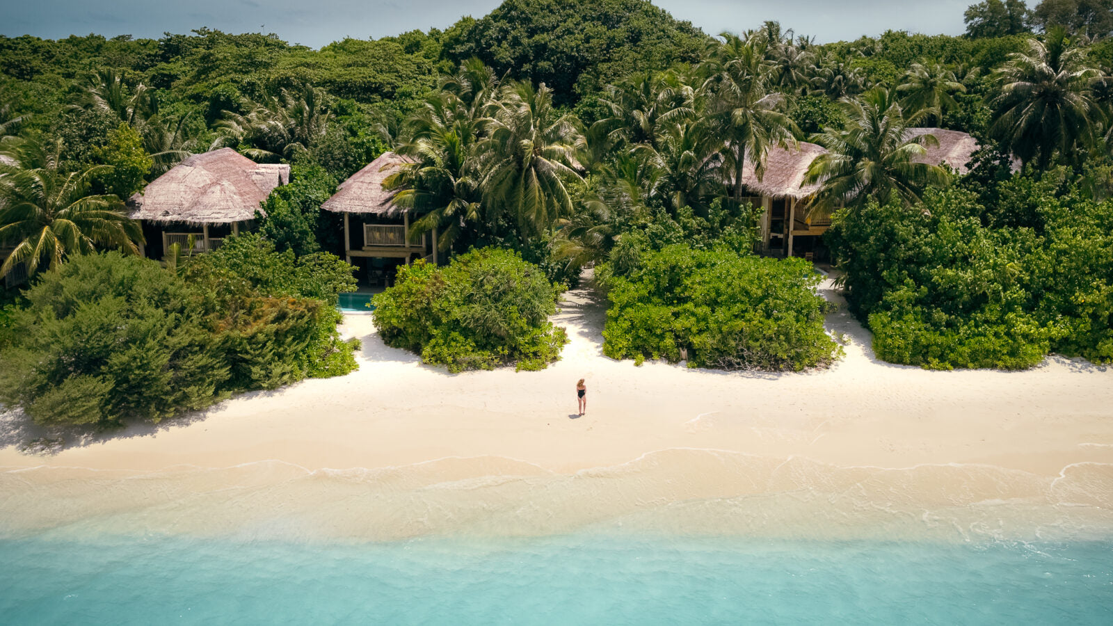 Aerial view of tropical beach with huts and a person walking on the sand.