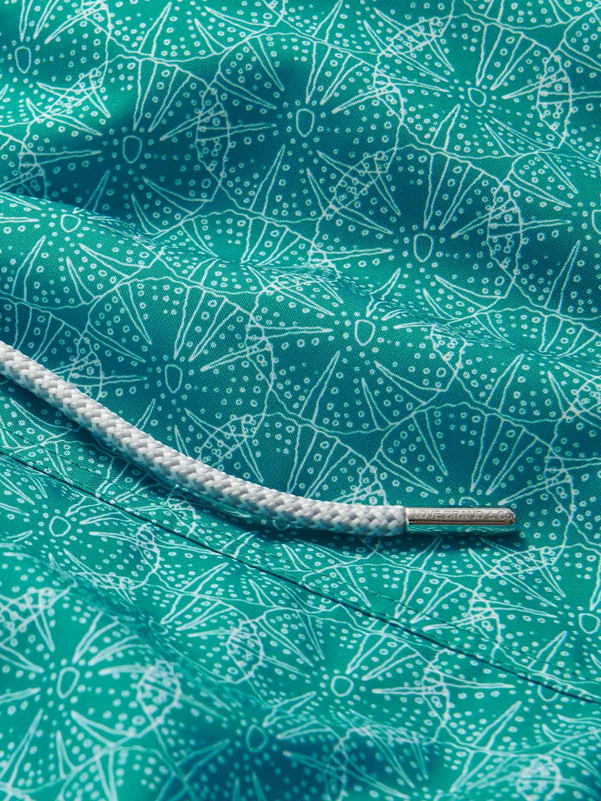 Close-up of Men’s Searching for Urchin Staniel Swim Shorts. Patterned swim shorts showing a sea urchin pattern on blue fabric and a white drawstring with a silver tip.
