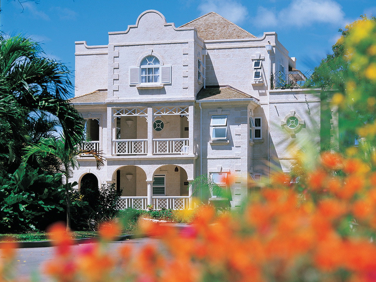 Large white building of the Coral Reef Club with a balcony, surrounded by greenery and flowers, under a blue sky.