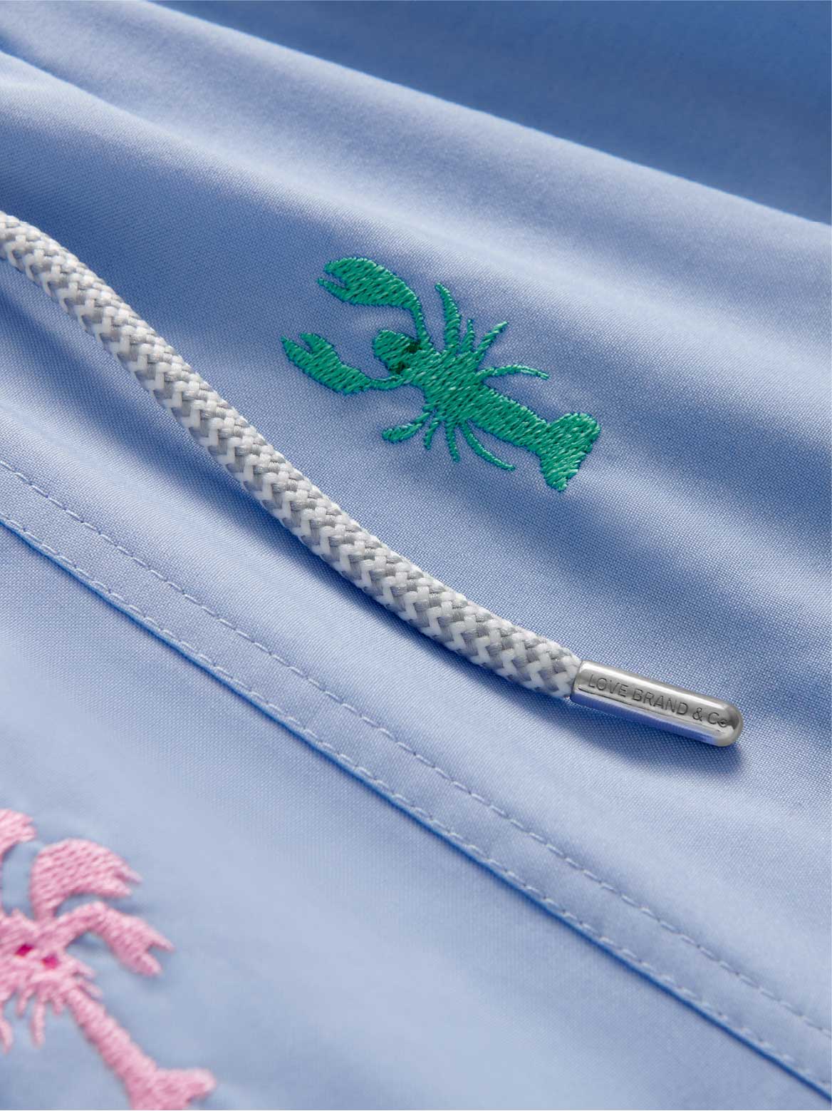 Close-up of a light blue fabric with embroidered green lobster and pink palm tree design.