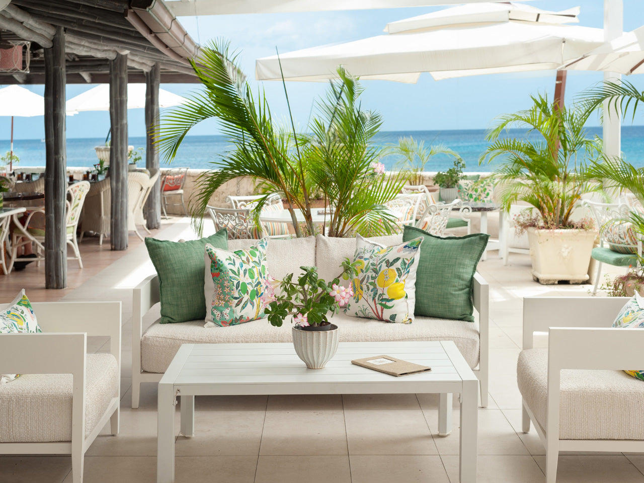 Outdoor seating area at Coral Reef Club with white furniture, green and patterned cushions, and a view of the ocean.