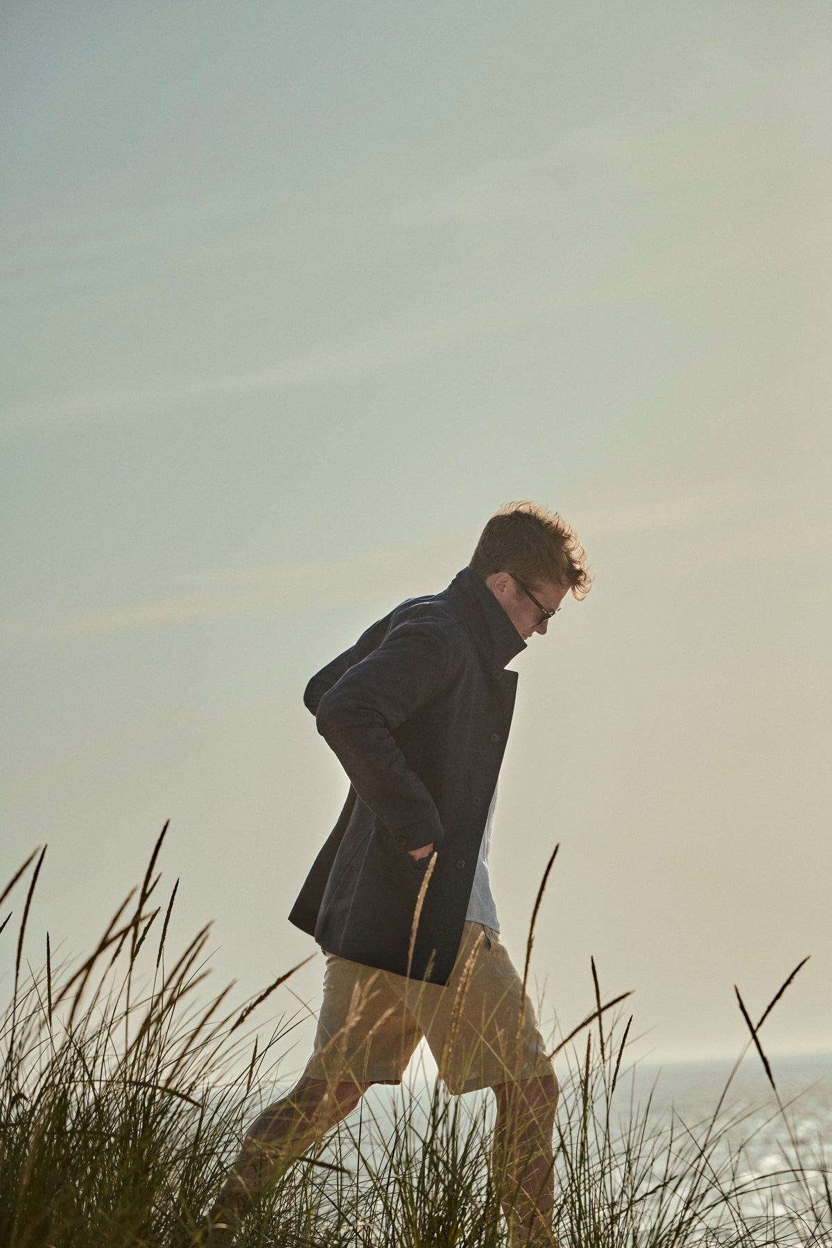 Ed Speleers wearing a dark navy coat, white t-shirt, and beige shorts walks through tall grass outdoors in warm golden-hour light.