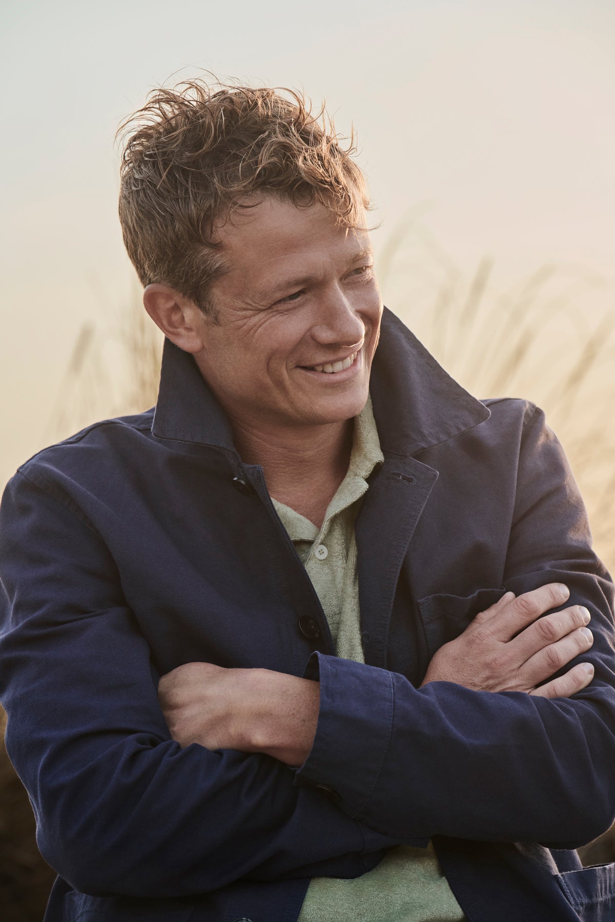 Ed Speleers smiling outdoors with arms crossed, wearing a dark navy jacket over a light green shirt, against a soft warm-toned blurred background.