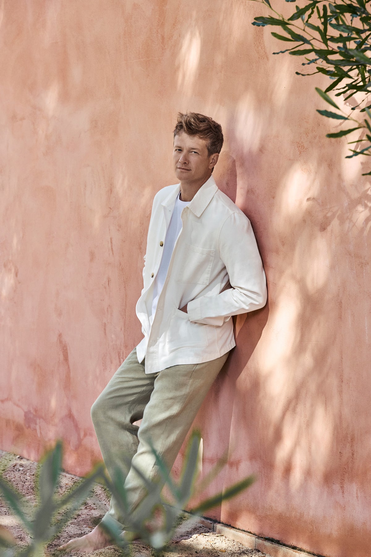 Ed Speleers wearing a white button-up cotton jacket and light olive green trousers leans against a textured terracotta wall outdoors.