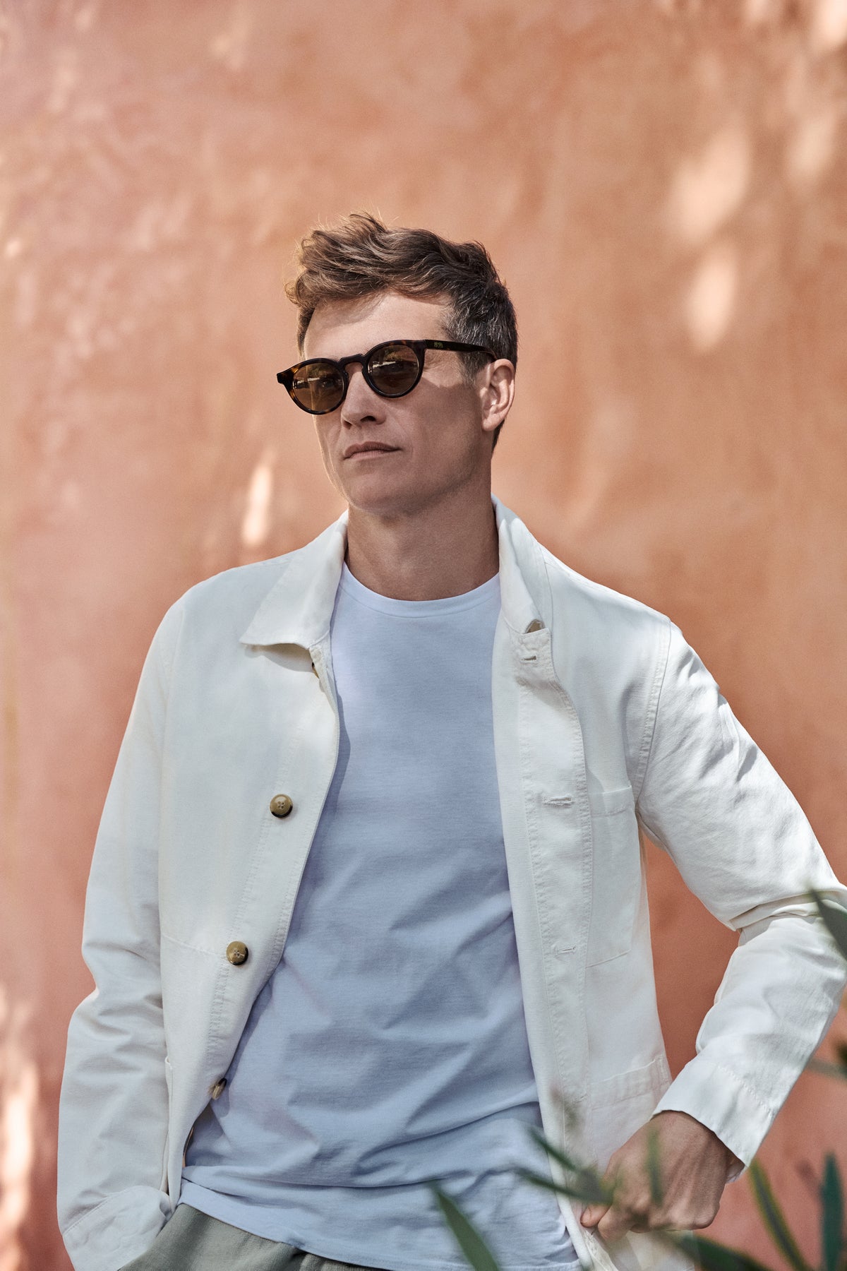 Ed Speleers wearing dark brown sunglasses, a light blue t-shirt, and a white jacket, standing against a textured terracotta wall.