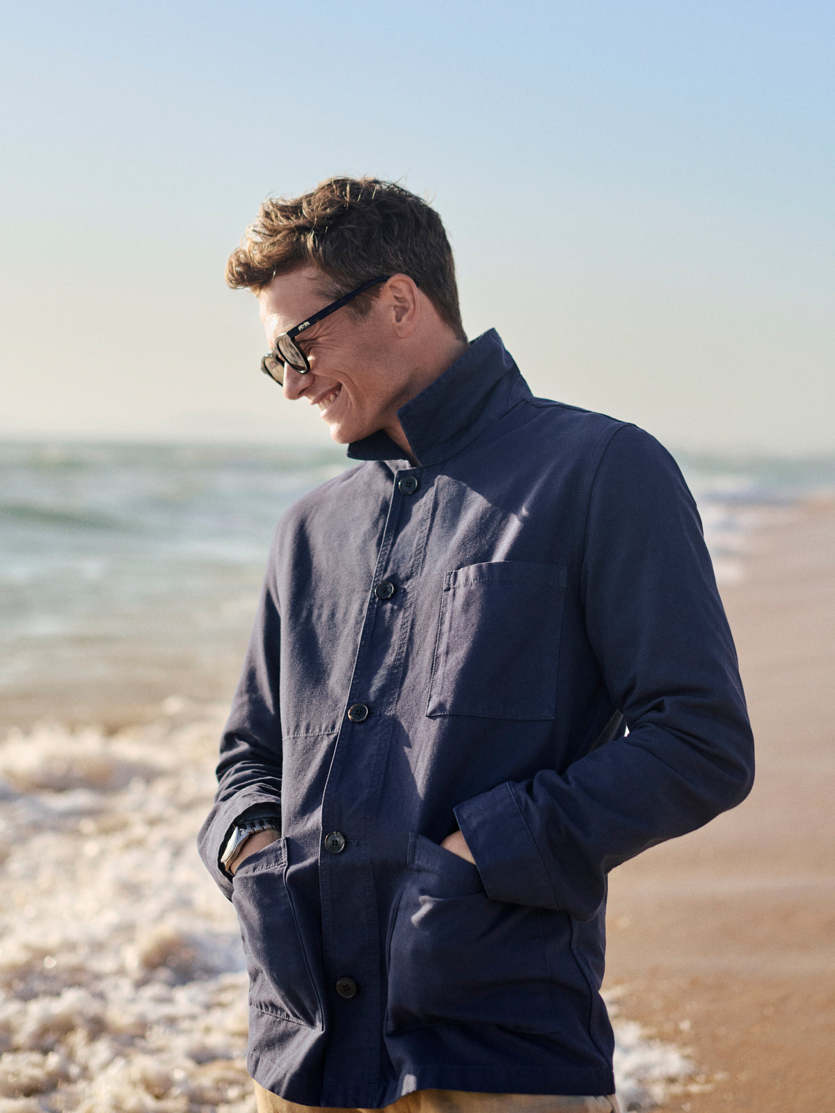 Ed Speleers wearing a navy blue button-up shirt jacket and sunglasses stands on a sandy beach with the ocean in the background.