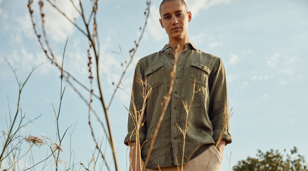 A man wearing an olive green button-down shirt and beige trousers standing outdoors in a field under a blue sky.