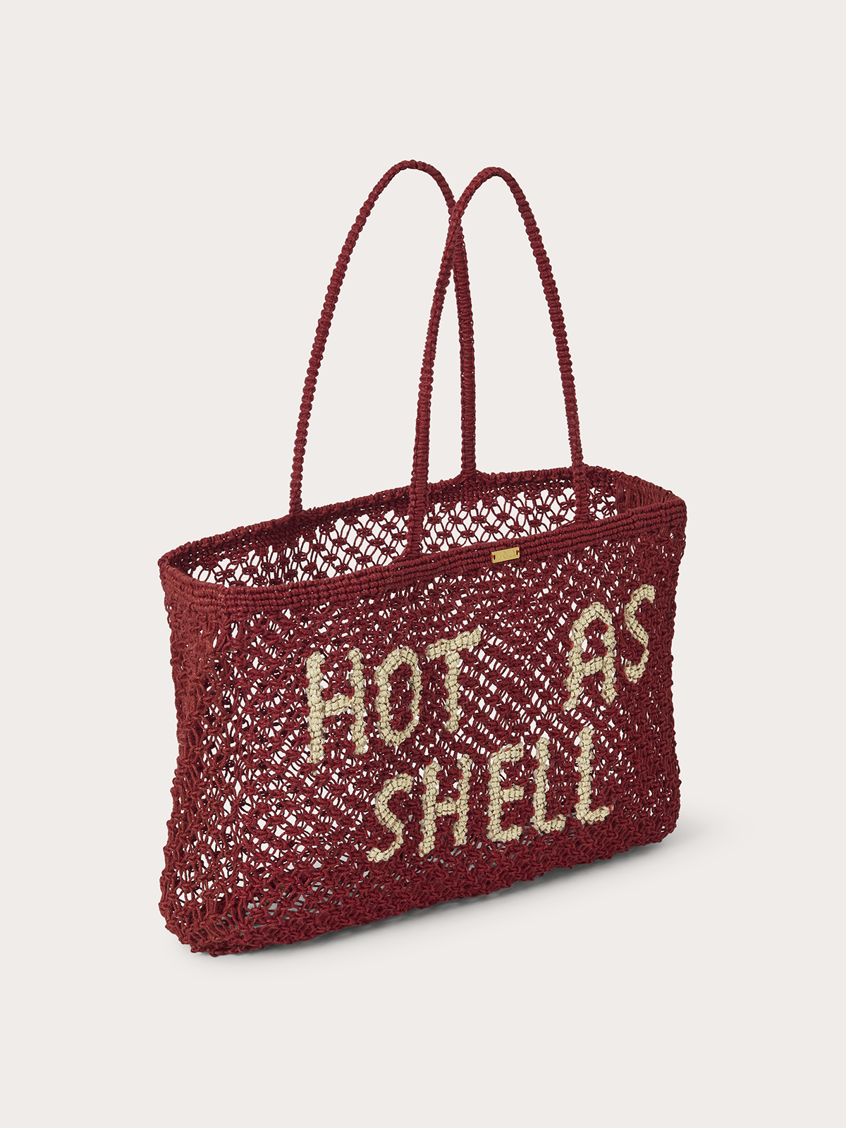 A red woven straw tote bag with two handles and the words 'HOT AS SHELL' woven in beige across the front, shown on a light grey background.