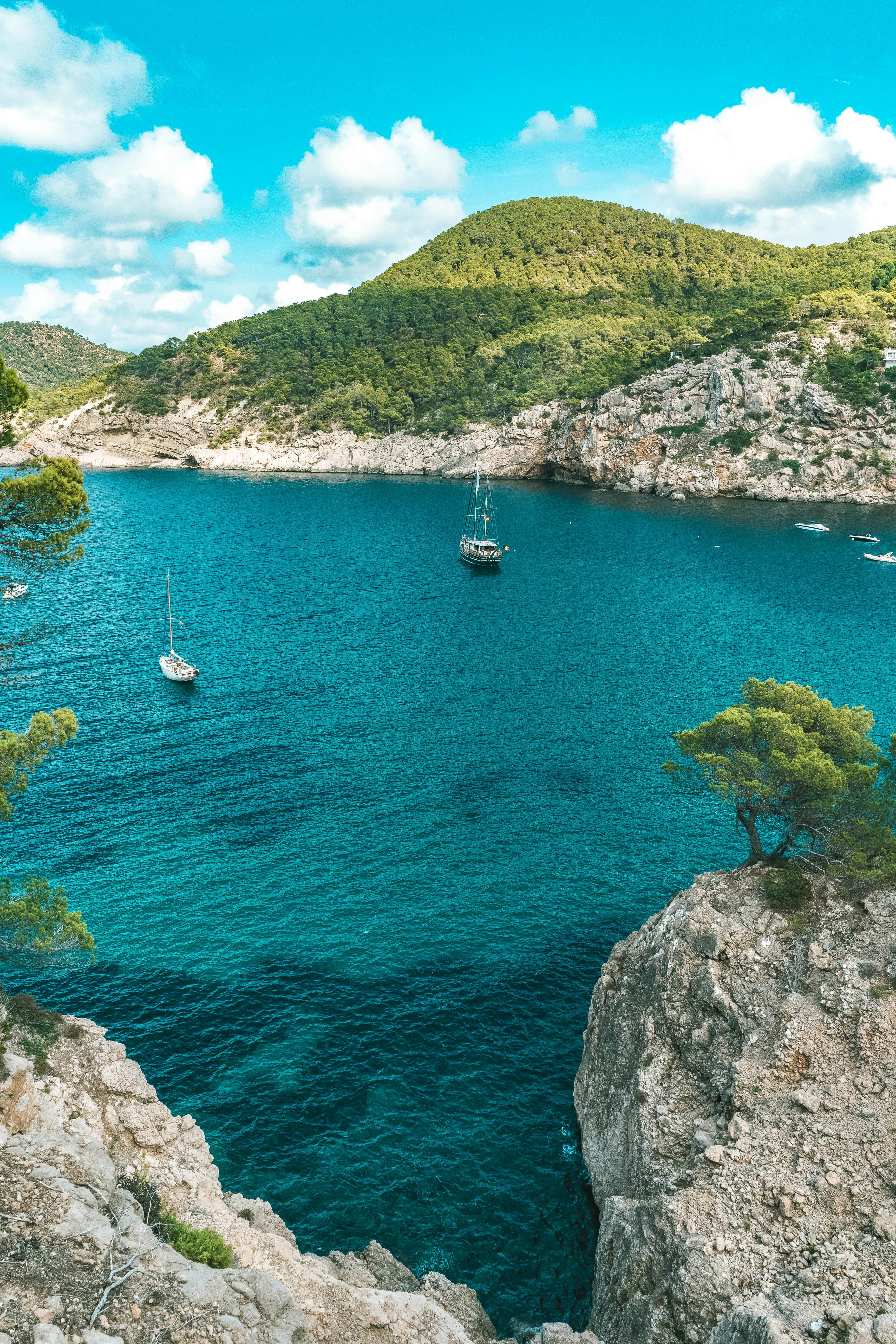 Two sailboats on a clear blue sea with a rocky coastline and green hills in the background.