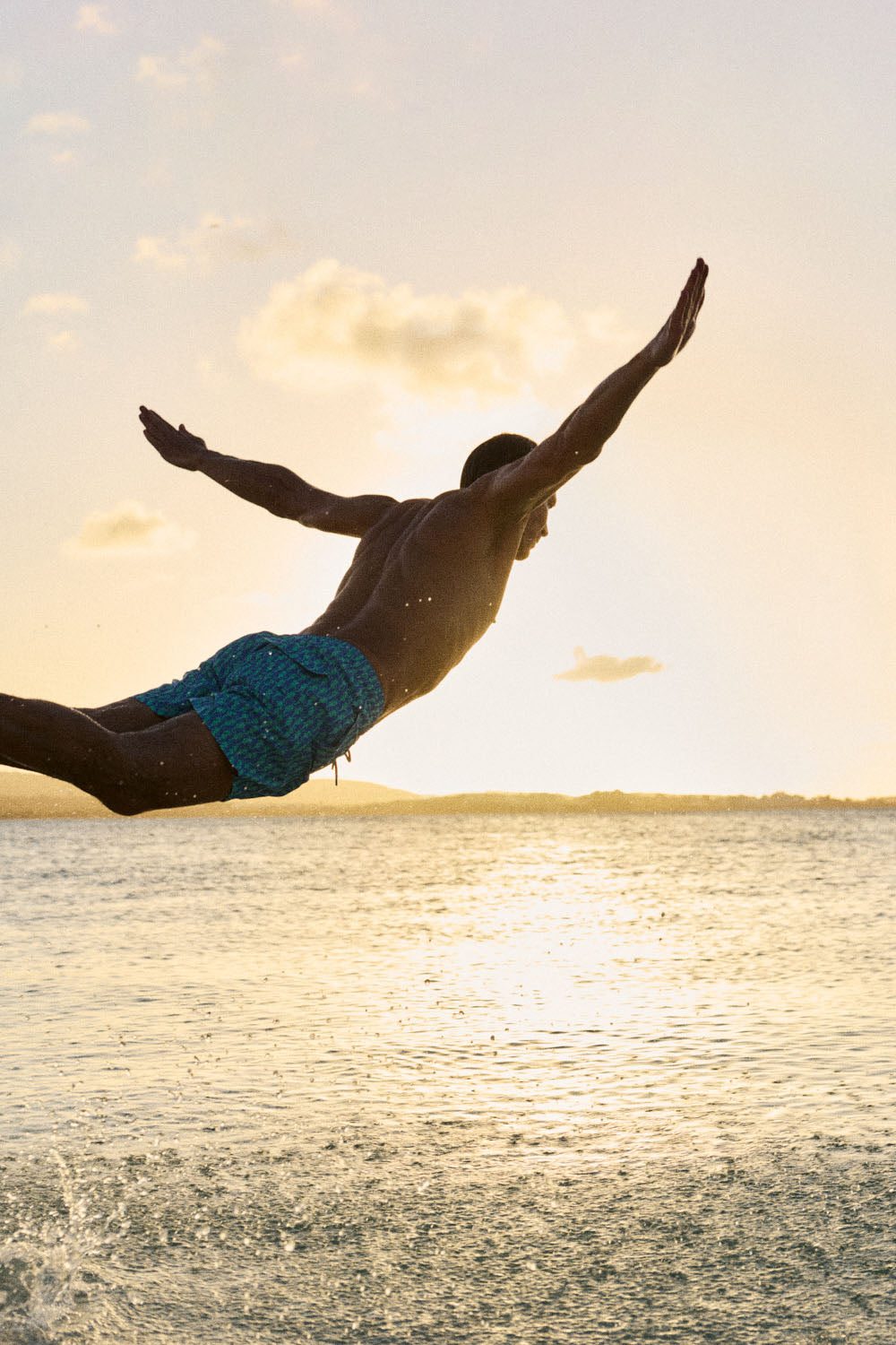 A man in blue patterned swim trunks jumps into the ocean from a diving board at sunset, silhouetted against a bright sky with clouds.