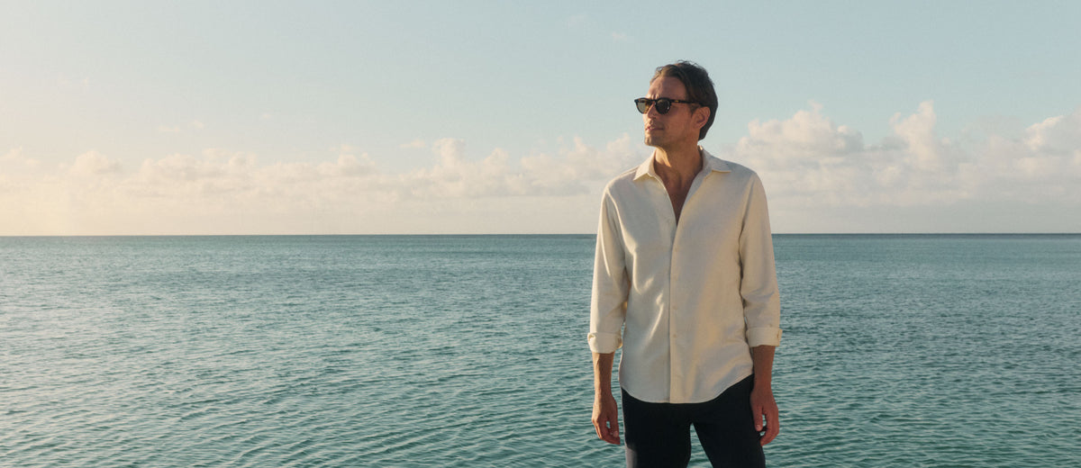 A man wearing a cream-colored long-sleeve button-up shirt and dark sunglasses stands on a beach with the ocean and a cloudy sky in the background.