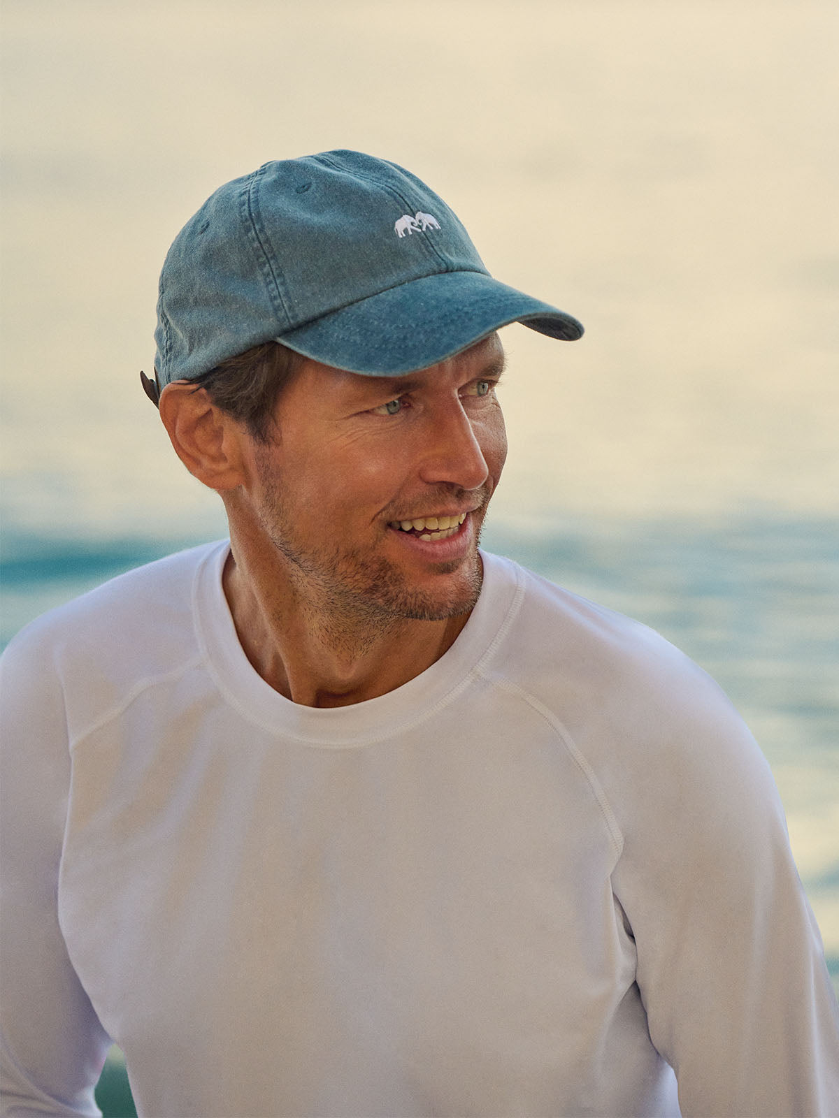 Man wearing a blue cap with a logo and a white rash vest, standing by the water.