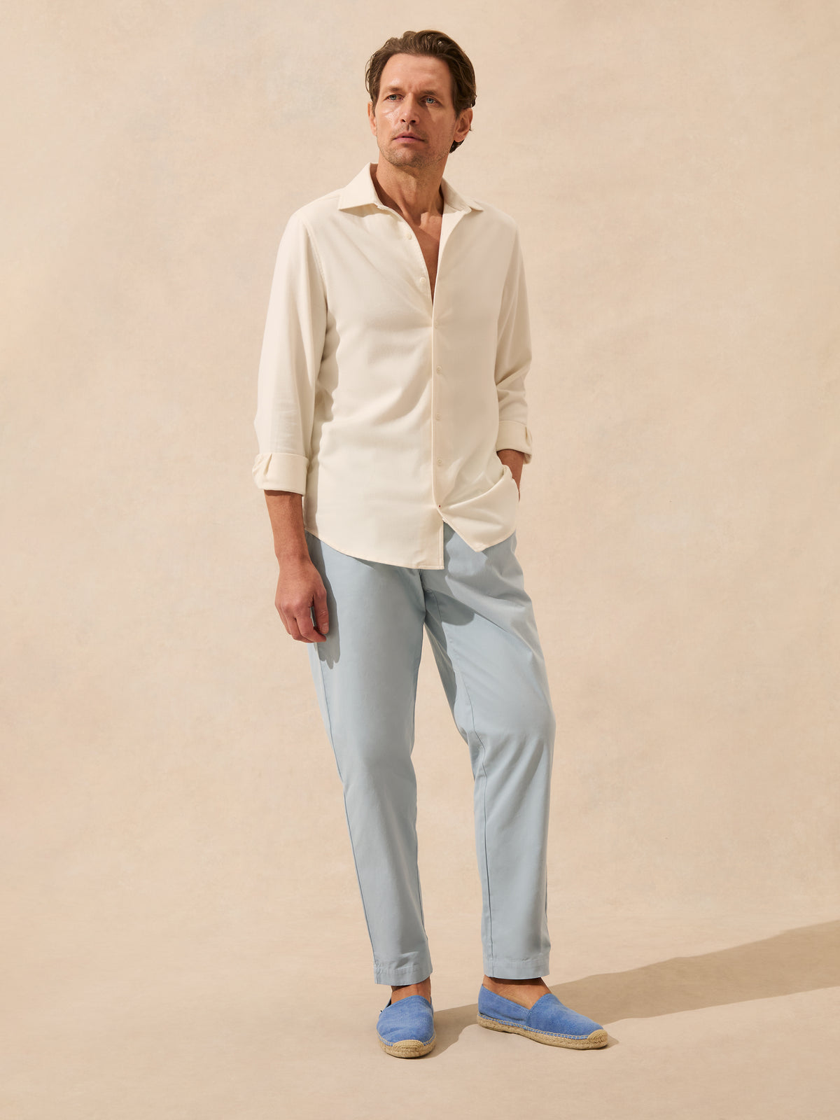 A man modelling a light cream long-sleeve button-down shirt, light blue trousers, and blue slip-on espadrille flats against a warm beige textured studio backdrop.