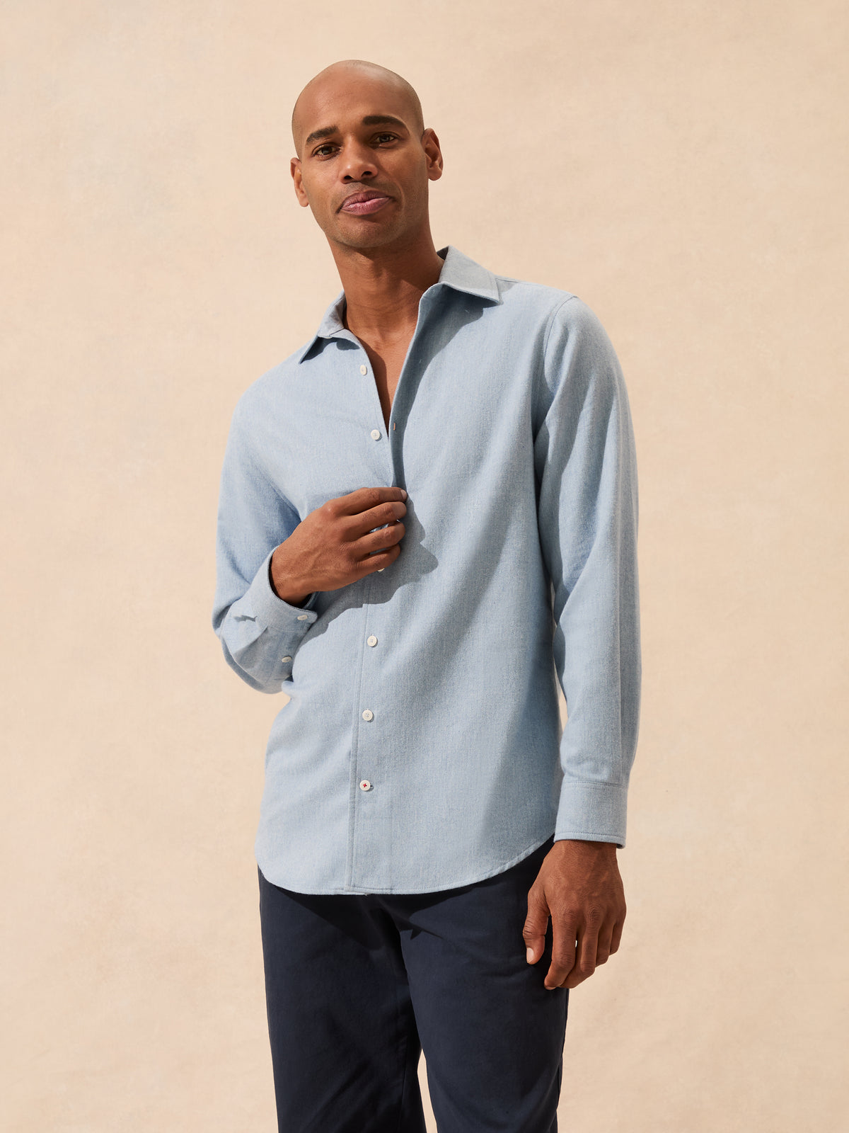 A man wearing a light blue long-sleeve button-up shirt and dark navy pants, standing against a warm beige textured background.