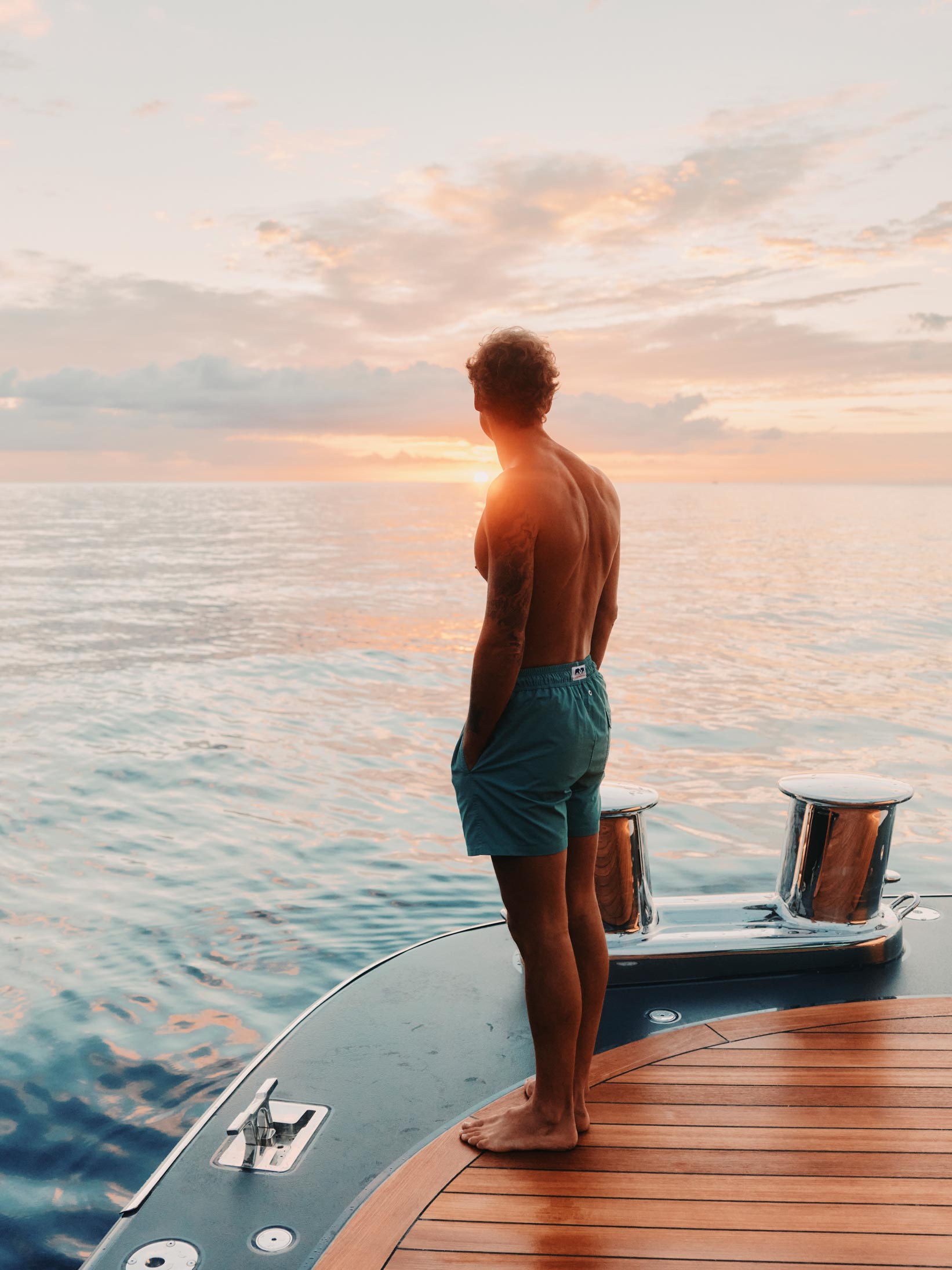 man wears swimwear london by beachwear brand love brand & co on a boat overlooking the sea at sunset.
