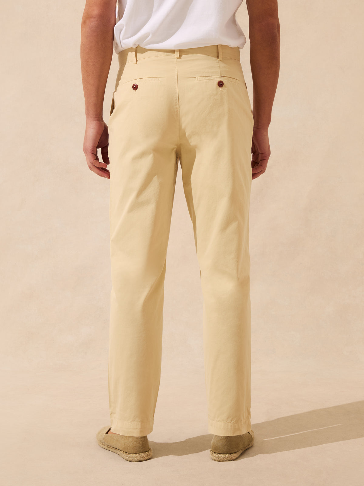 A man viewed from the back wearing light beige chino pants, a white t-shirt, and brown espadrille sandals, standing against a warm beige textured studio backdrop.