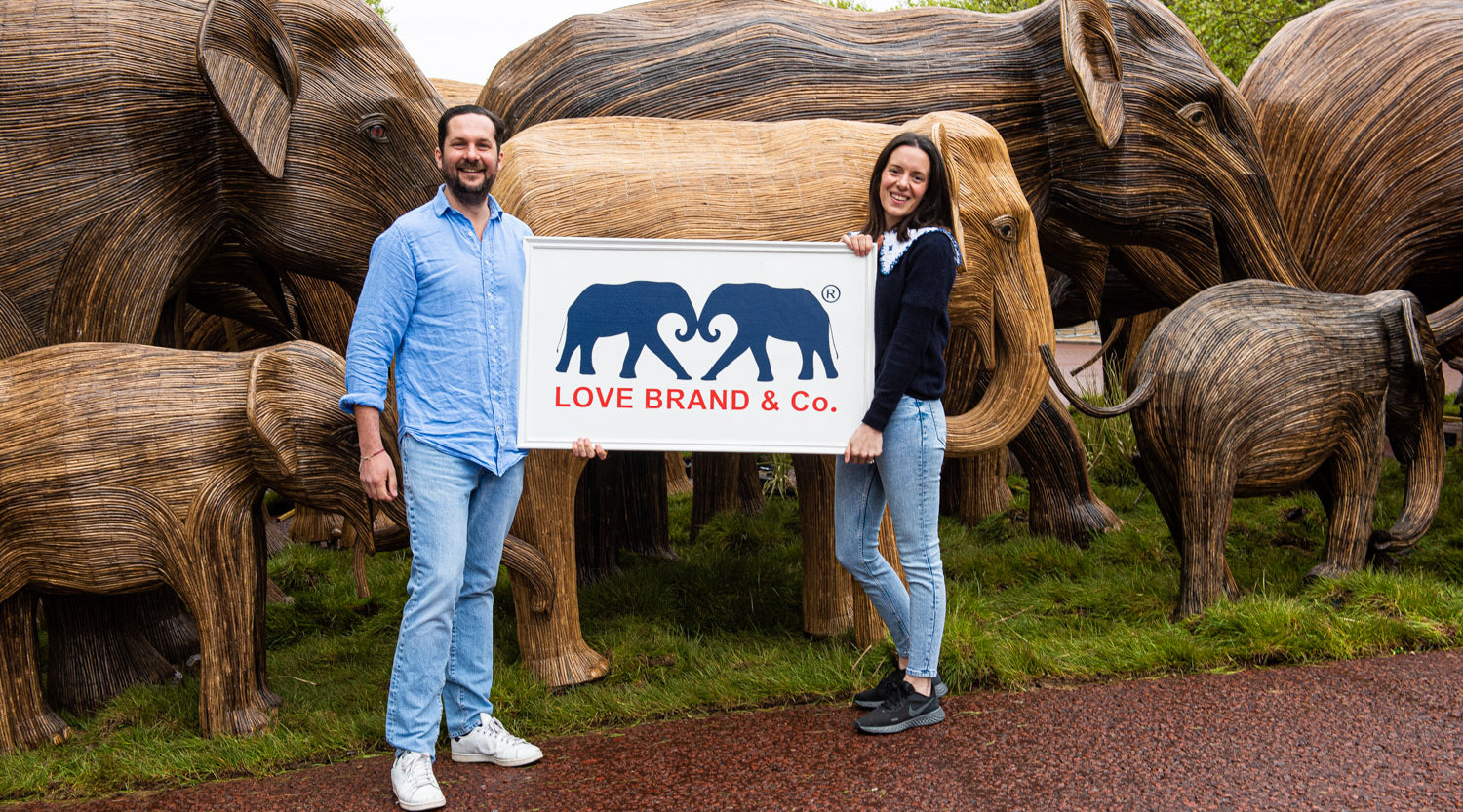 Two people holding a sign with 'Love Brand & Co.' in front of wooden elephant sculptures.
