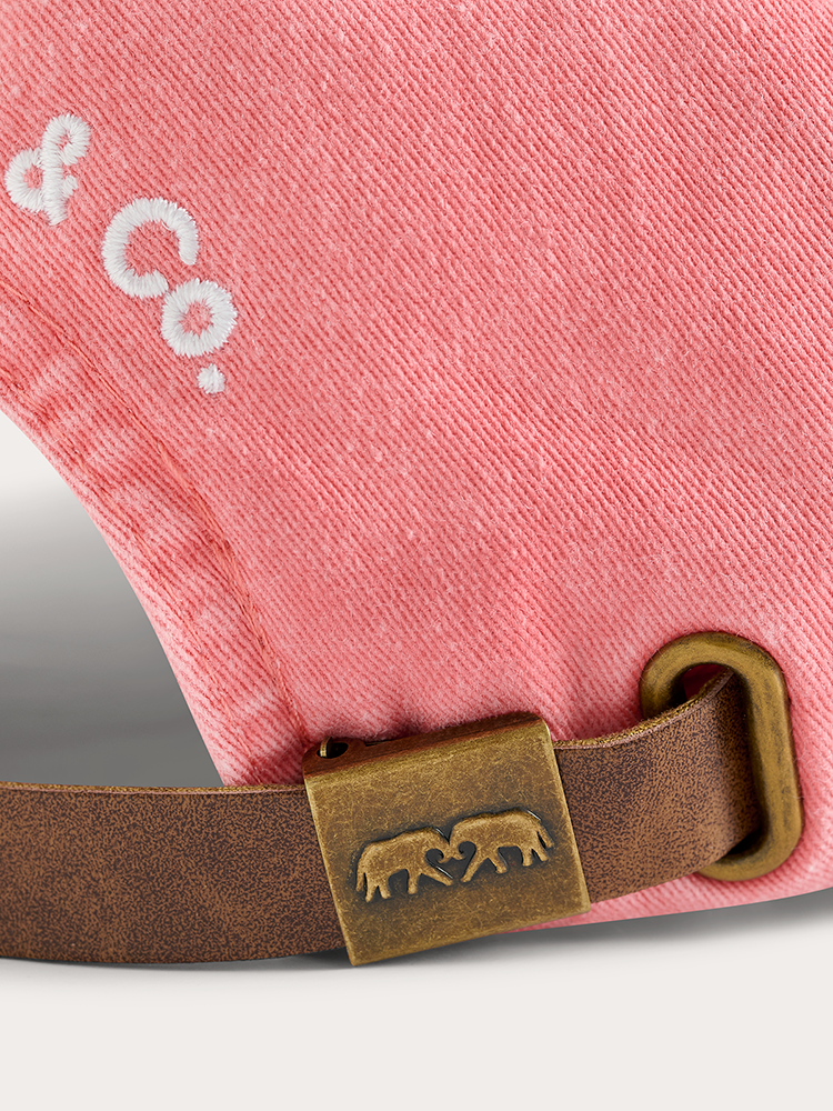 Close-up of a pink denim cap with a brown leather strap and metal buckle, featuring a brand logo.