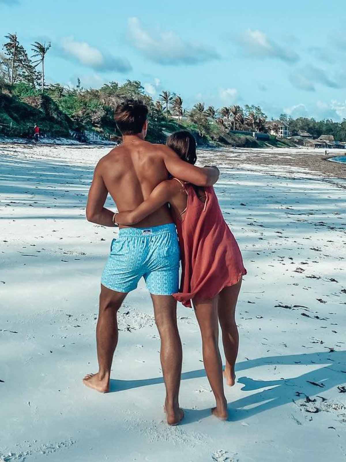 man wears blue swim shorts by resort wear uk brand love brand & co as he walks across the beach.
