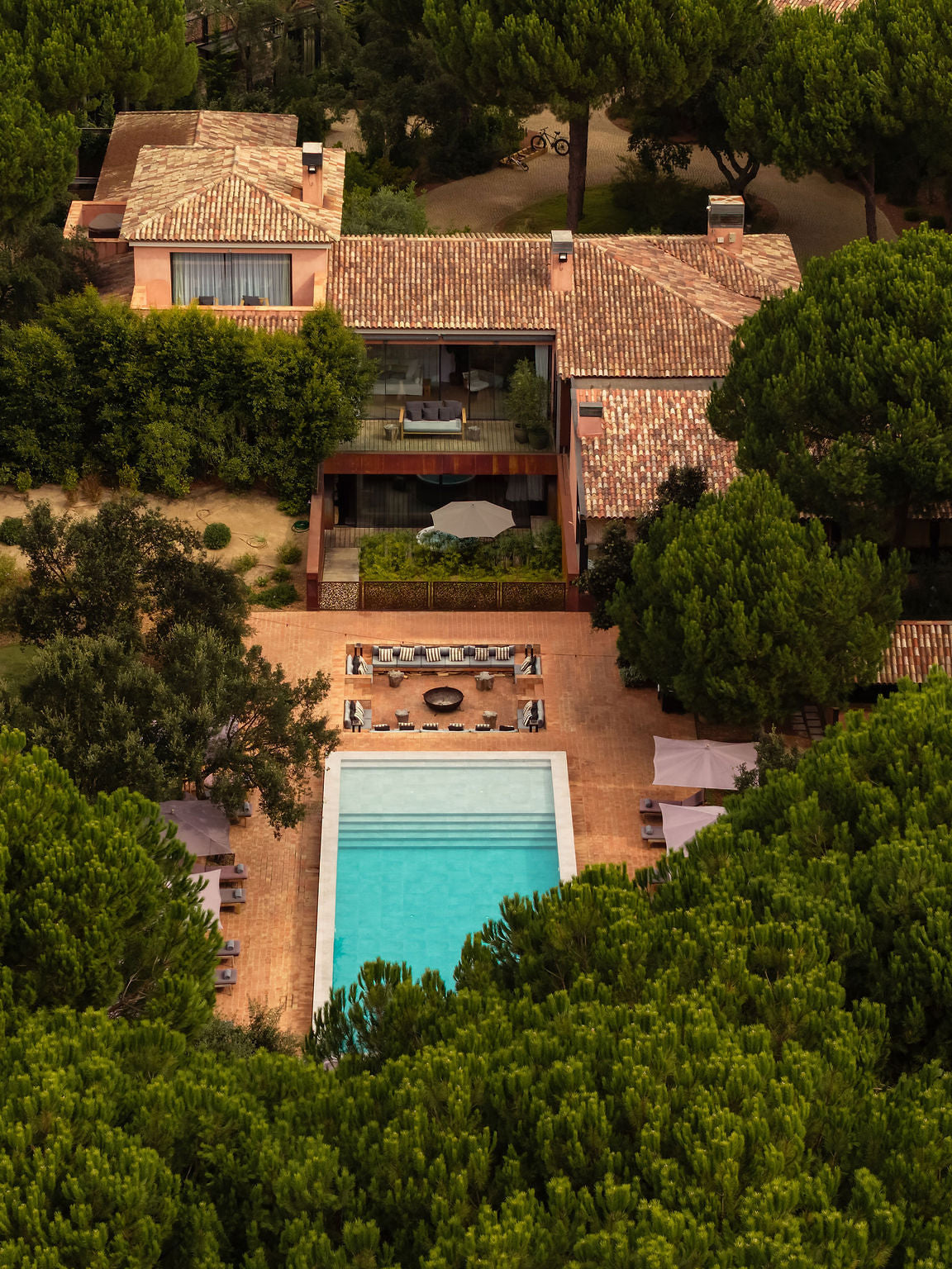 Aerial view of a luxury villa with a terracotta tiled roof, a rectangular turquoise swimming pool, and a patio area, surrounded by dense green trees.