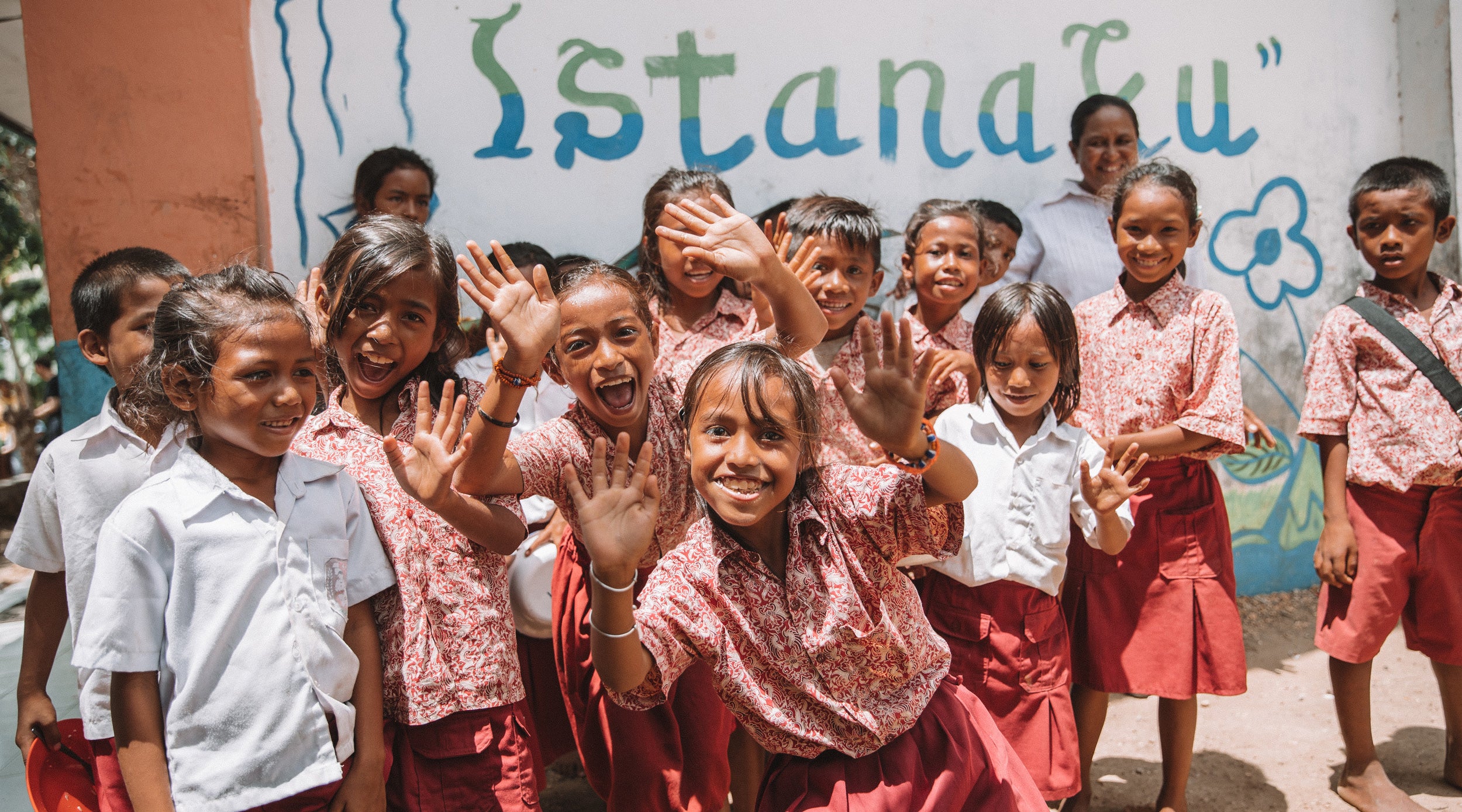 Children waving at the camera with a colorful wall in the background
