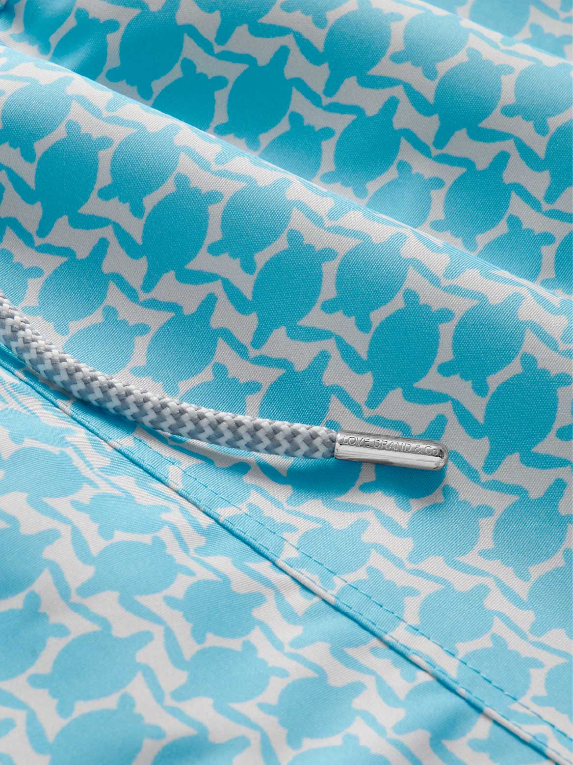 Detailed view of Love Brand mens swim shorts. Close-up of a drawstring on top of a blue printed turtle pattern.