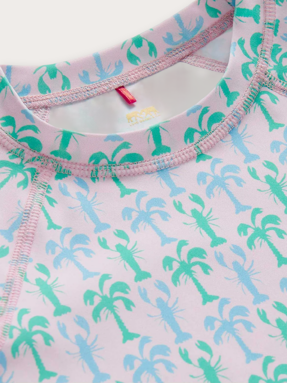 Close-up of a children's long-sleeve rash vest with a light pink base, featuring an allover print of green and light blue palm trees and lobsters