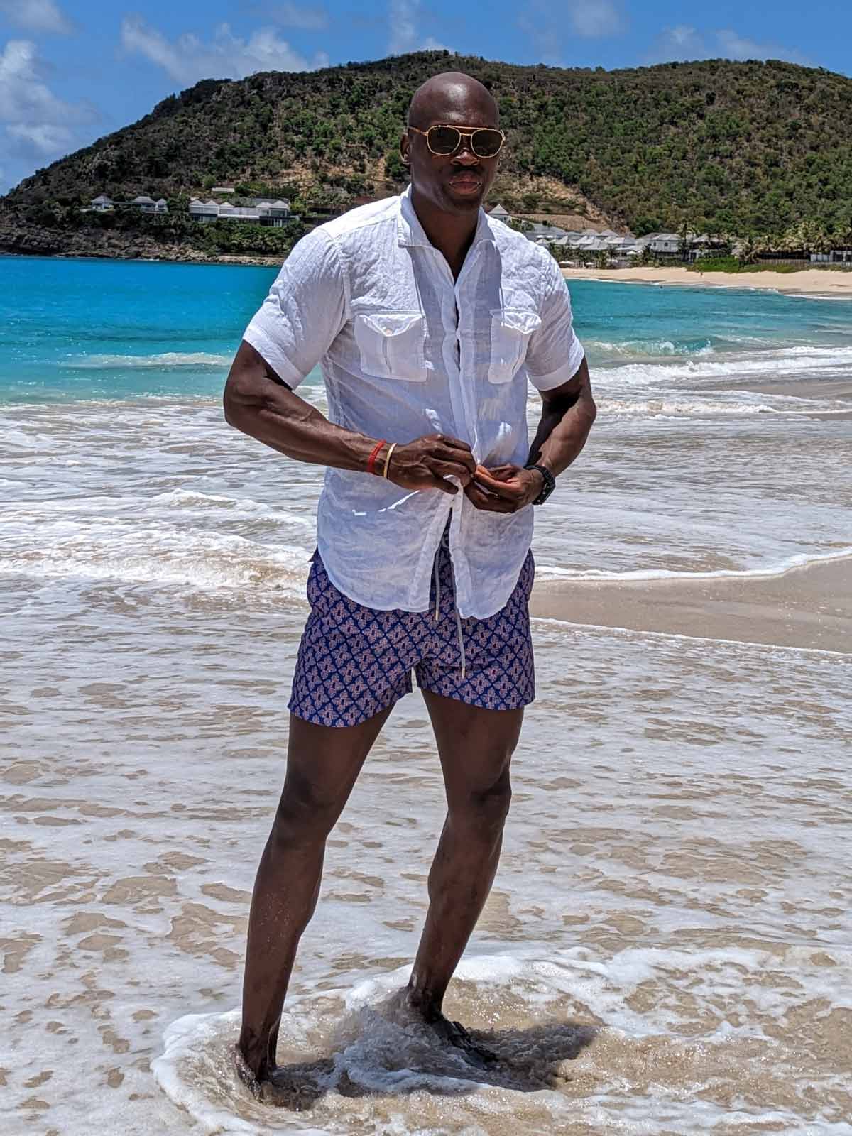 man wears white linen shirt and swim shorts by luxury swimwear brands uk as he stands in the ocean.