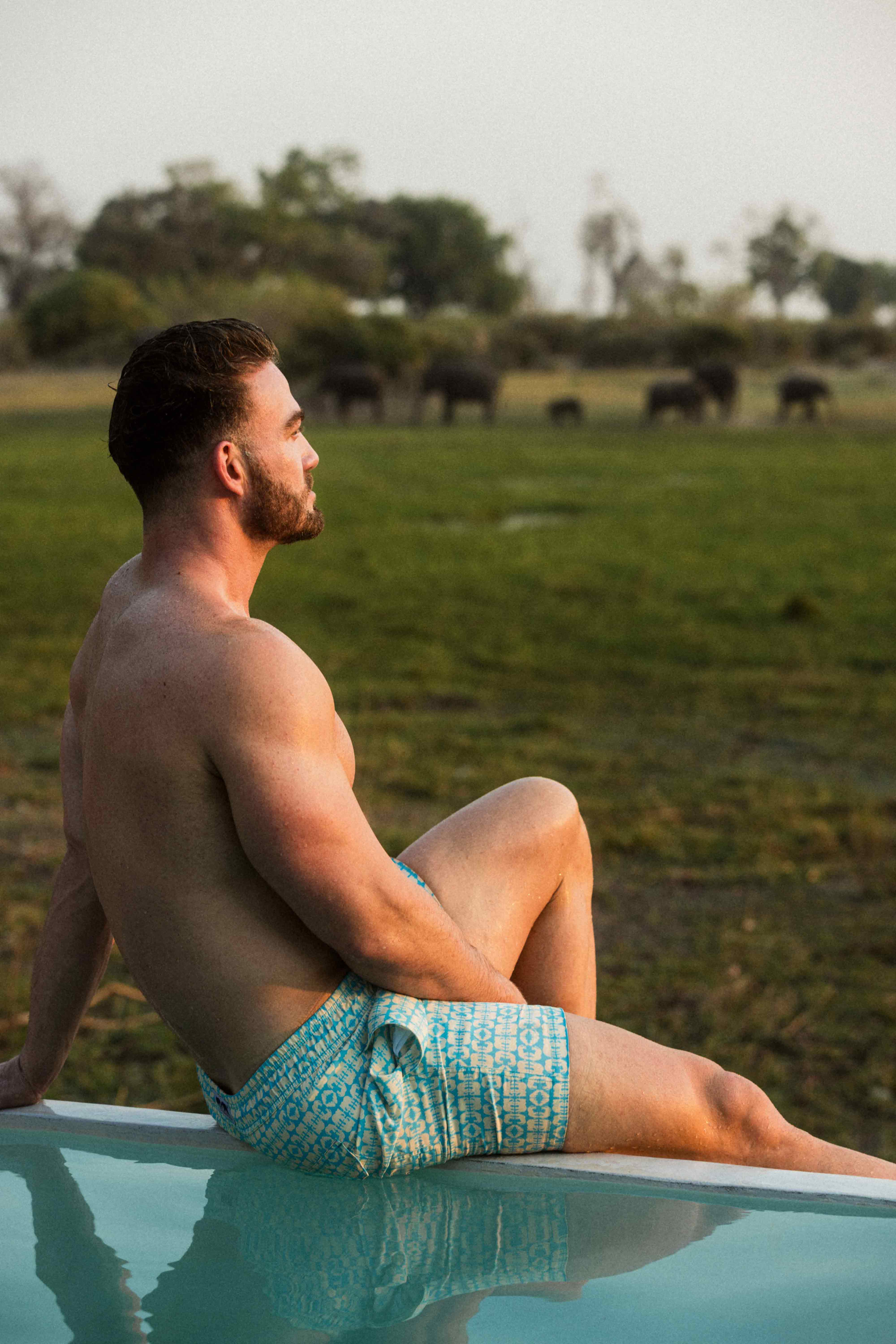 Man in blue swim shorts sitting by a pool with elephants in the background