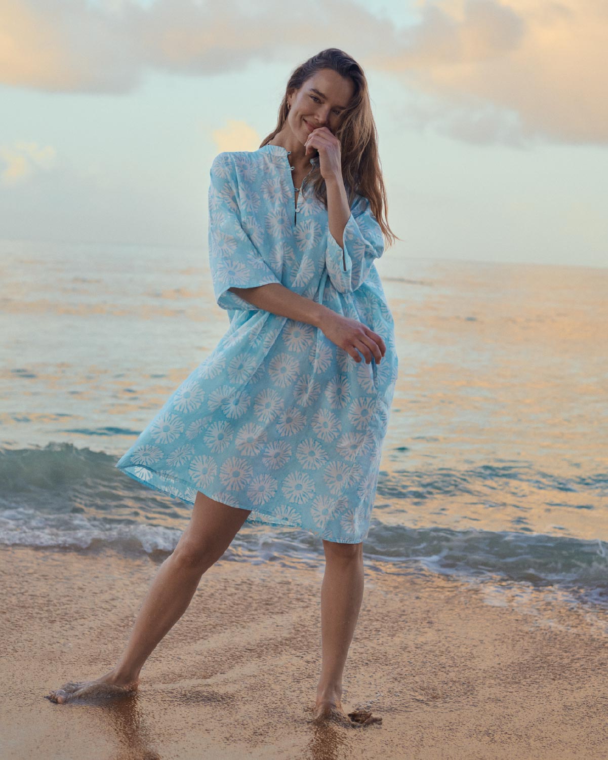 Woman in a light blue floral dress standing on a beach at sunset.