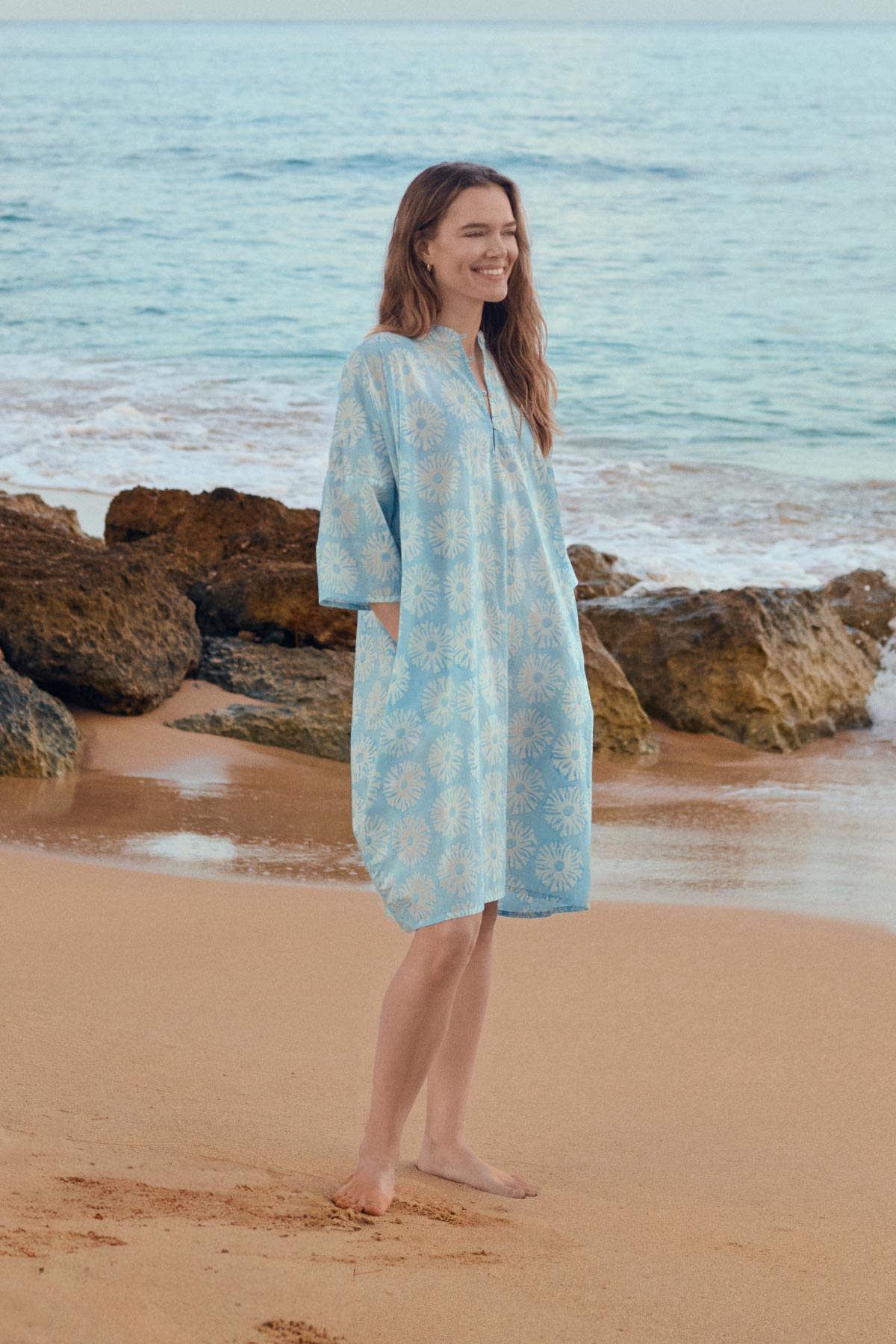 Woman in a light blue floral dress standing on a beach with ocean and rocks in the background