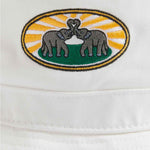 White cotton twill bucket hat with vibrant woven patch of two elephants with intertwined trunks, set against a background of sun rays and greenery.