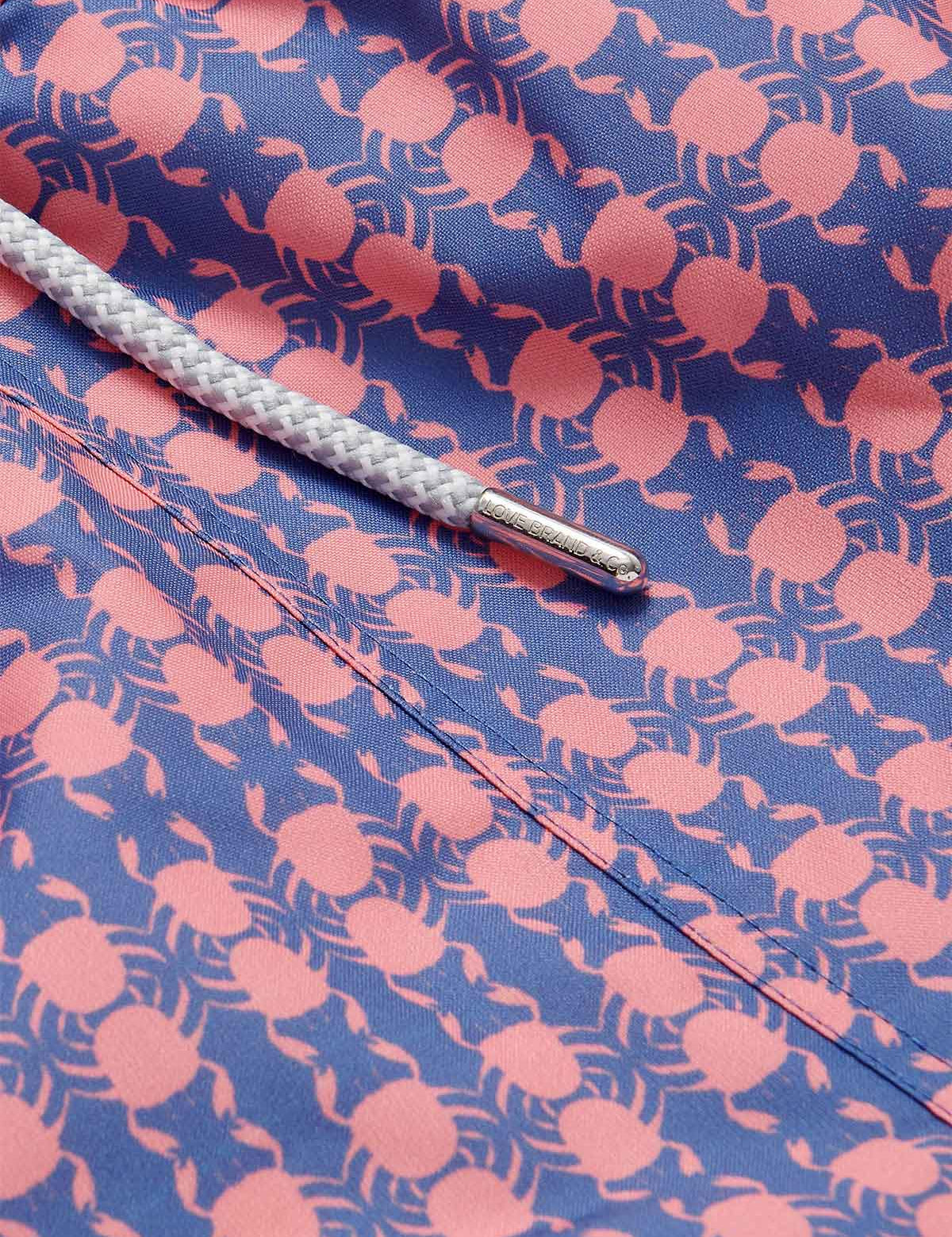 Close-up image of Men's Crab Pool Staniel Swim Shorts. Patterned swim shorts featuring a pattern of pink crabs on a deep blue base with a white drawstring.