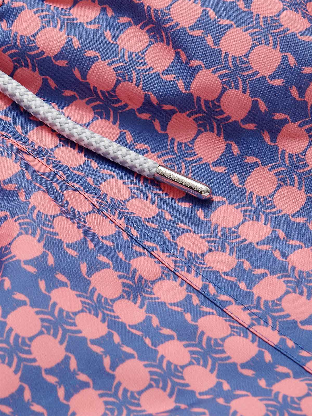 Close-up image of Men's Crab Pool Staniel Swim Shorts. Patterned swim shorts featuring a pattern of pink crabs on a deep blue base with a white drawstring.