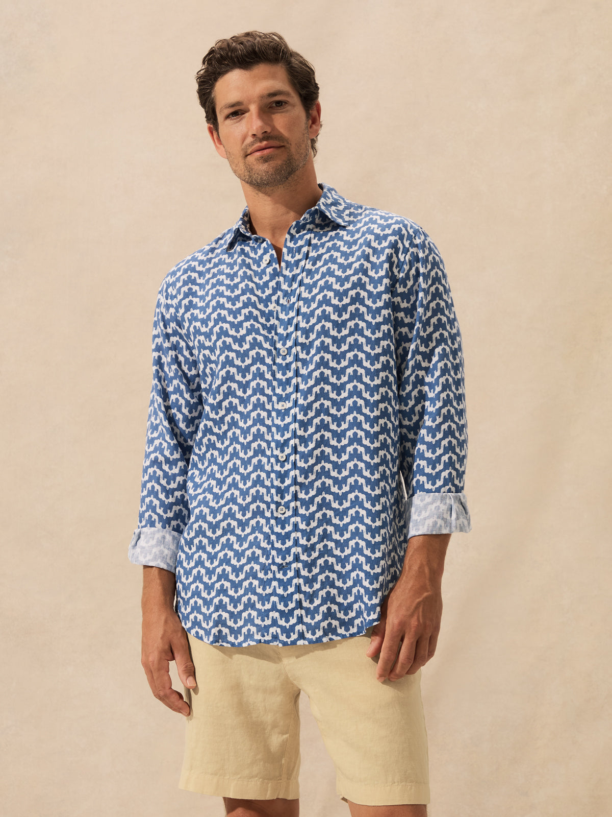 A man wearing a blue and white patterned long-sleeve button-up shirt with beige linen shorts, standing against a textured beige background.