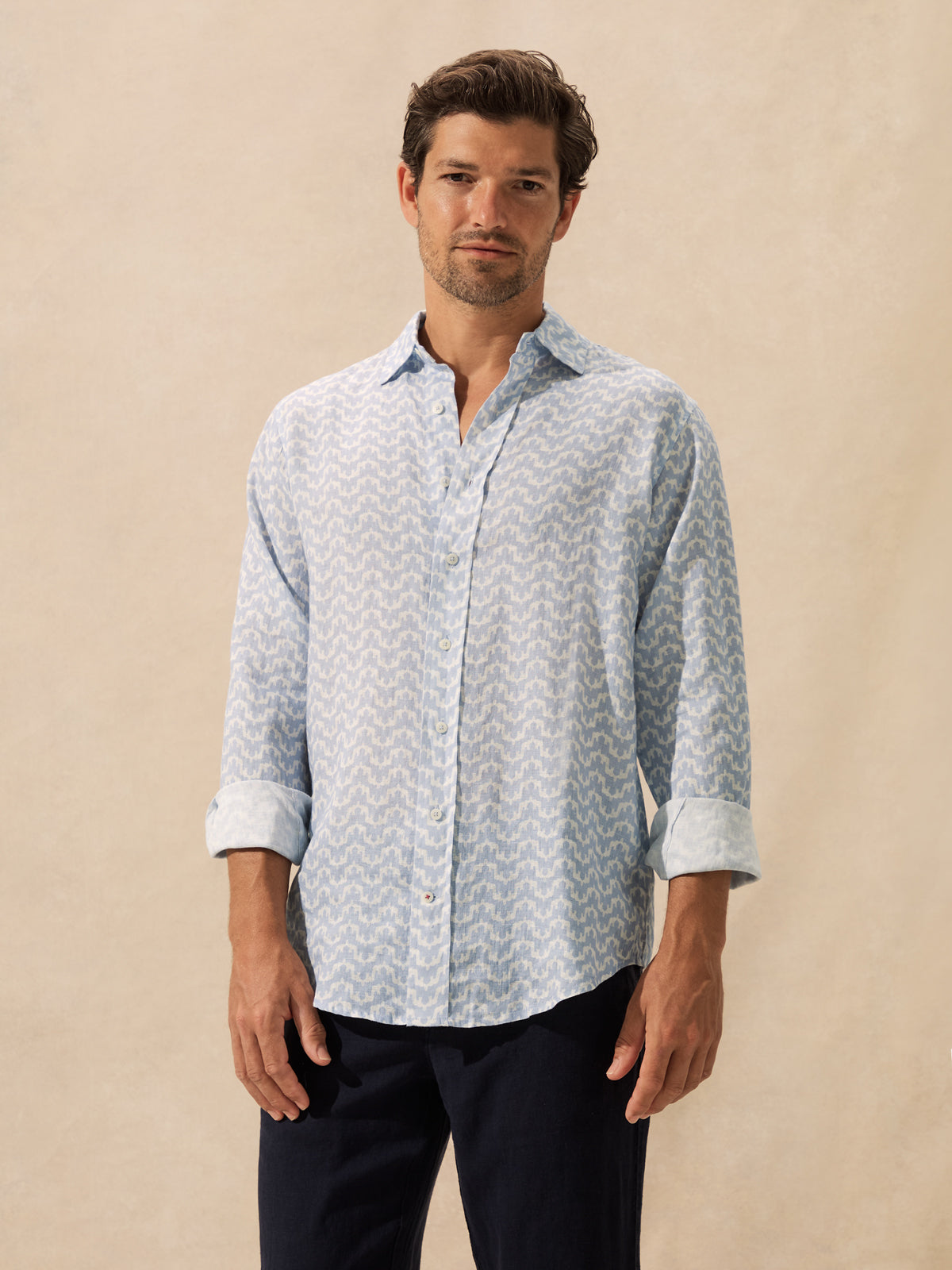 A man wearing a light blue and white patterned button-down shirt standing against a neutral beige background.
