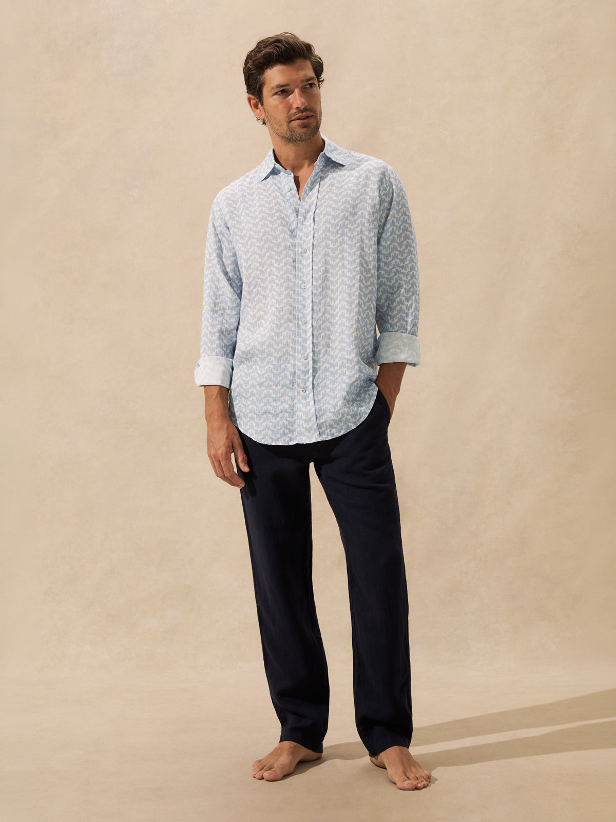 A man stands barefoot against a warm beige studio backdrop, wearing a light blue and white patterned long-sleeve button-up shirt with dark navy pants.