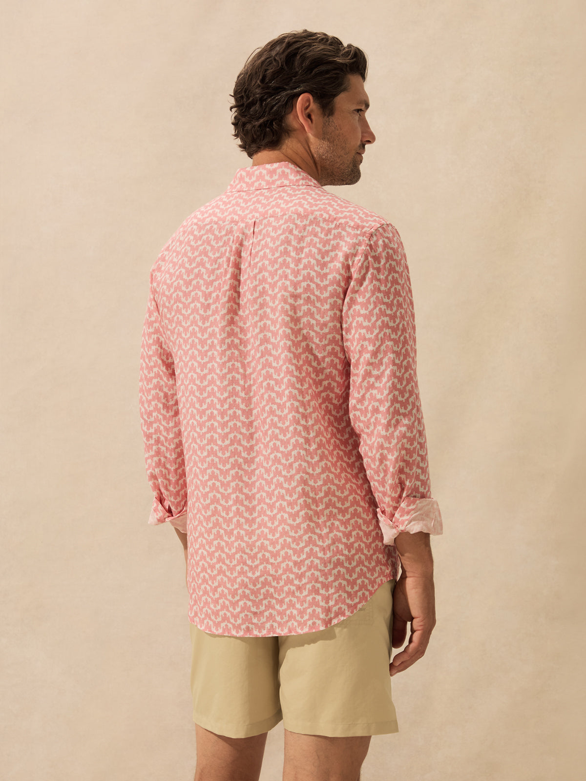 A man shown from the back wearing a pink and white patterned long-sleeve button-up shirt standing against a textured beige background.