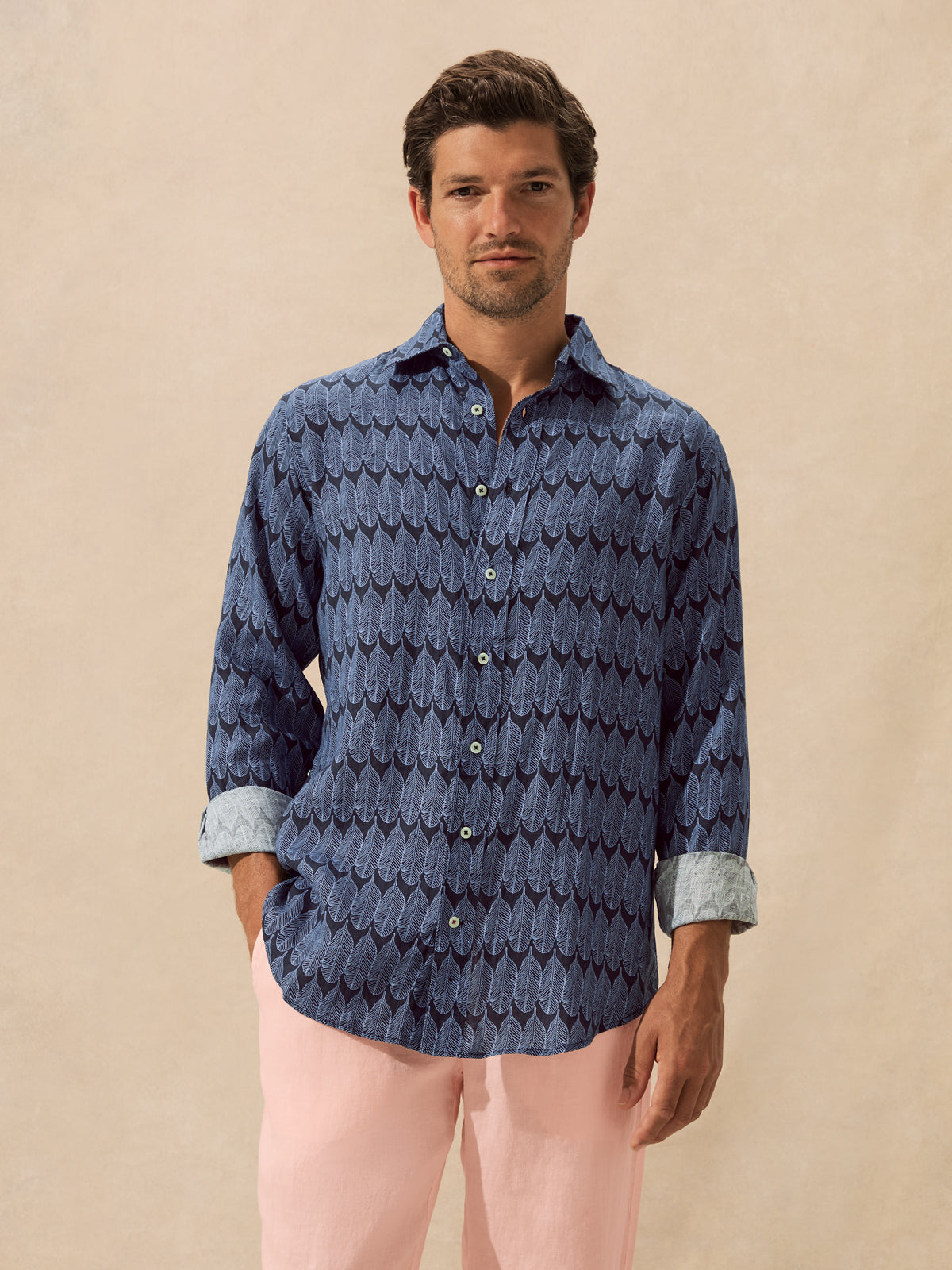 A man wearing a blue long-sleeve button-up shirt with a dark repeating leaf pattern and light cuffs, paired with light pink pants, standing against a plain beige backdrop.